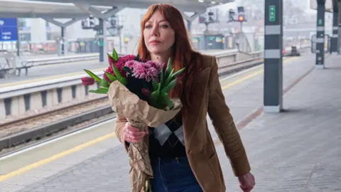The image shows a woman walking on a train platform holding a bouquet of flowers.
