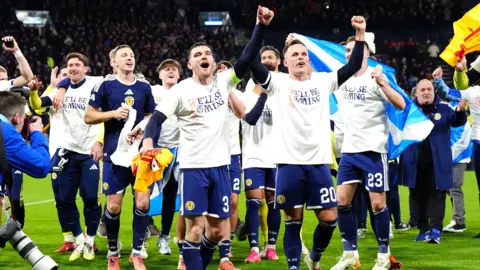 PA Media Scotland players celebrate after reaching the World Cup. Several have their arms raised joyfully while others salute the fans. Most of the team are wearing T-shirts saying We'll Be Coming.