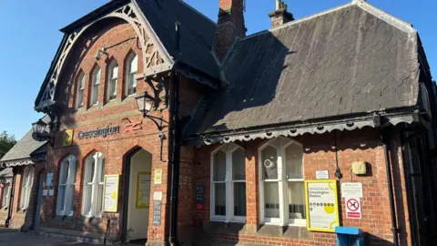 BBC/Claire Hamilton A pretty red brick train station with eight widows, all with white frames. The historic building has lamps over the entrance, and Cressington in black lettering alongside the National Rail and Merseyrail symbols. 