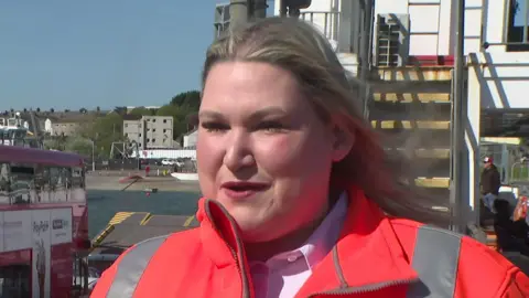 Kellyann Bastos, ferry manager for Tamar Crossings. She is wearing a red high-vis jacket and has long straight blonde hair. She has lipstick on. She is stood in front of a flight of steps. A red double decker bus is to the left of the photo.
