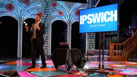 MP Jack Abbott, in a suit and tie, is giving an address to an audience off-screen, while on stage at the New Wolsey theatre in Ipswich. Next to him is a TV screen with the words City of Culture Ipswich in white lettering on a sky blue background.