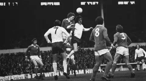 Getty Images A black and white image showing two footballers contesting a header on the pitch as other players look on.