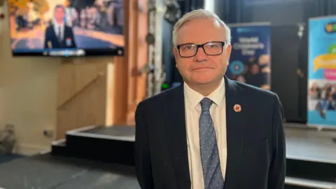 BBC A man wearing a suit looking towards the camera. He is wearing glasses. 