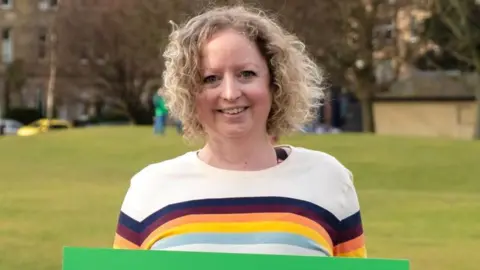 A woman with blonde curly hair smiles at the camera. She is standing in a park. 