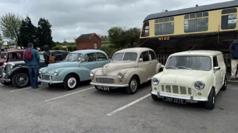 A row of classic British cars including two Morris Minors and a Mini parked in a car park. Some people are admiring the car at the end of the row. 