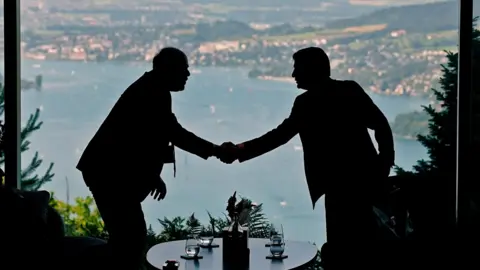Two figures in silhouette shake hands with a lake visible behind them, at talks in Switzerland between the US and Iran