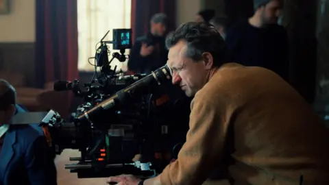 Lol Crawley, who has short greying hair and is wearing a brown jumper, looking through the lens of a large black camera which is pointed at something off the left side of the shot. In the background is a window with red curtains, and various film crew members dressed in black.