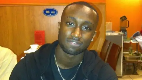 Julian Cole when he was well, looking at the camera and smiling. He wears a black t shirt and hoodie and silver chain around his neck. 