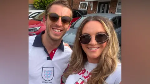 A man and a woman stood next to each other wearing England football shirts and sunglasses. They are smiling.
