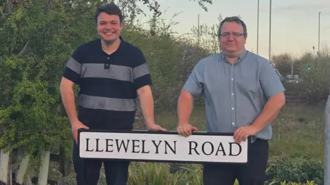 Daniel Adams Councillors Daniel Adams and Jake Chandler standing behind a street sign with the name 'Llewelyn Road' with grass and bushes in the background.