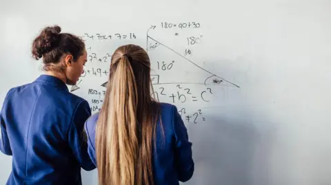 Getty Images Two students wearing navy blue blazers face away from the camera as they write algebra on a whiteboard. The student on the left has her brown curly hair up in a bun, and her classmate on the right has long light brown hair that is half tied up.