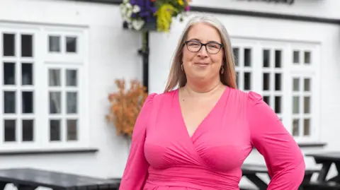 Nicki Cameron Hannah is wearing a bright pink long sleeved top, she has light blonde hair and dark glasses. Behind her is a white building with black benches outside and foliage on the drain pipe