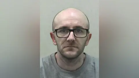 Mugshot of McConville. He is completely bald and has a stubble beard. He is wearing black-framed glasses and a grey sweatshirt