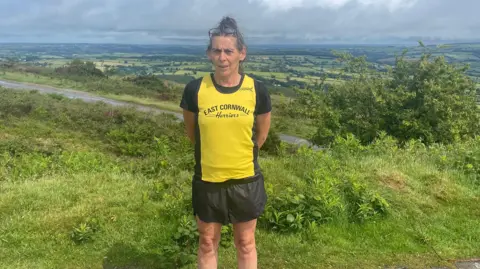 A woman in a yellow jersey that says "East Cornwall Harriers" and in black shorts. She has her hair in a bun and glasses on her head and is looking directly at the camera. Dartmoor spreads out behind her, there are green fields. There are grey clouds in the sky.