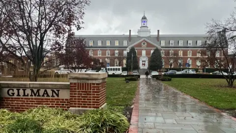 A large brick building can be seen in the background with a brick gate that says Gilman in the foreground. It is rainy and the sky is grey.