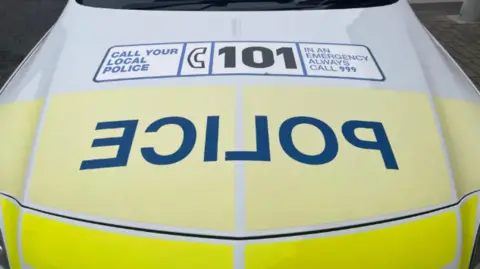 The front of a yellow police car which has the words 'POLICE' on the front. Above it is the words 'CALL YOUR LOCAL POLICE' with '101' in the centre and 'IN AN EMERGENCY ALWAYS CALL 999'.