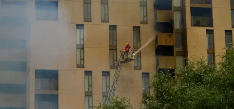 Atilla Vida A firefighter on an aerial ladder sprays a hose reel onto a tower block whose balconies are blackened from heat and smoke. 