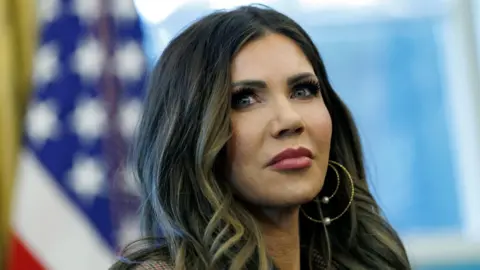 EPA Kristi Noem's face pictured in front of a blurry background with the stars of the US flag to the left of the image. 