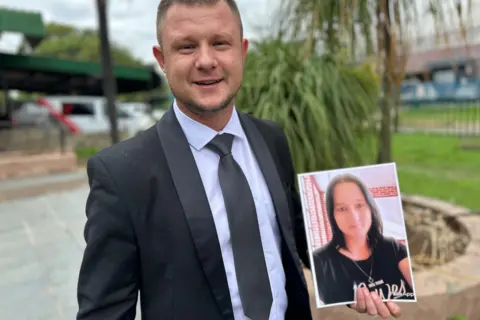 BBC Jason Pretorius, in a suit, is seen holding an image of his mother, Charlene