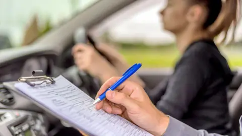A driving instructor holds a pen to a clipboard in a car whilst a young woman takes her test in the driving seat
