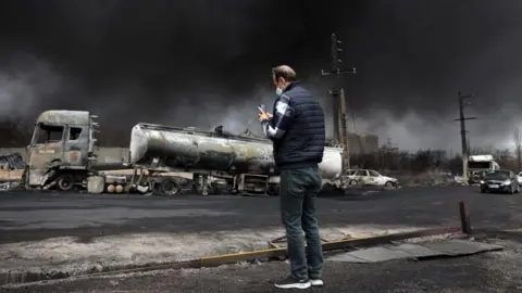 EPA Smoke rises and vehicles lie ruined after an attack on an oil refinery in Iran, with a man standing in the foreground taking a picture f the wreckage 