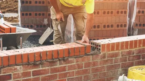 Getty Images A builder is laying bricks. The wall is up to his knees, with mortar squeezing out of the joints. In the background, to the left is a wheel barrow with more red bricks in it, with stacks of additional bricks to the right behind the builder