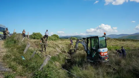 PA Media A red and grey mini digger is removing pieces of grass from the ground. Around the digger a collection of people are watching, some are wearing camouflage clothing while others are dressed in navy. The land is cordoned off by a wire fence. At the edge of the land a vehicle is sitting. In the background is a row of mountains.