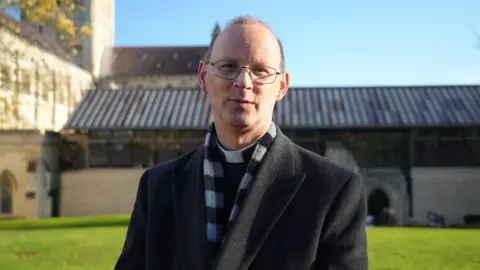 Shaun Whitmore/BBC A head and shoulders photo of the Dean of Norwich Cathedral, the Very Revd Dr Andrew Braddock. He is wearing a grey coat, grey and black scarf and clerical collar. He has short grey hair and wears thin rimmed glasses.