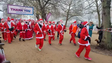 Keech Hospice Care A large number of people running in a Santa Run, wearing red and white Santa suits with hats and trainers on. Spectators are taking photos, and there are dogs, too. A river is in the distance, along with a white suspension bridge. The sky is misty and there trees, with no leaves on, along the river