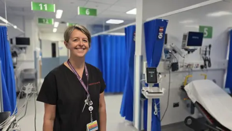 A woman with short blond hair wearing a black nursing gown. She is in a hospital room with cubicles with blue curtains. There is a gurney to her right. 