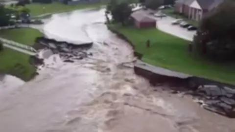 A road can be seen broken up, with brown water flowing through the centre of it.