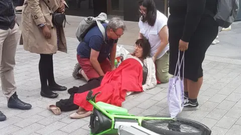 A woman is lying on the ground covered in a red blanket. A lime bike is on its side in front of her.