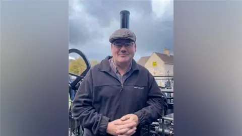 Ironbridge Gorge Museum Trust A man wearing a brown flat cap, glasses and a black zip fleece is standing in front of a small black steam engine with a tall chimney.