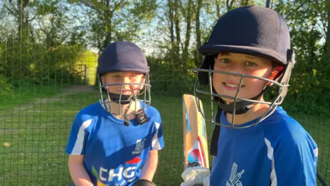 Two primary school aged boys, who play for West and Middle Chinnock Cricket club, are photographed in the new cricket nets with a cricket bat and ball. 