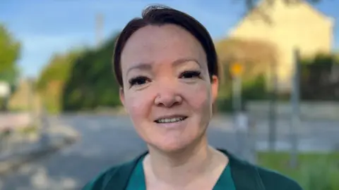 Kayleigh McCormack is smiling and looking directly at the camera with her dark hair pulled back from her face. She has brown eyes and her nose pierced, dark eye make-up and is wearing a green work uniform. There is a village road blurred in the background.