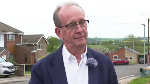 A man with short hair, wearing glasses and a blue suit with a white hair. In the background are several red brick houses on an estate, a road and cars.