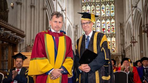 Two men wearing red and golden graduation robes, smiling with their hands closed in front of them, with standing in large hall with stained glass windows and other university officials behind them.