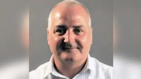 A head and shoulders shot of Sexton who is wearing an open neck white shirt. He is smiling and has very short grey hair. 