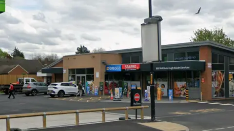 A Google Street View screenshot of the shops at Roseberry Service Station in Middlesbrough. The low, flat-roofed building has signs for Greggs and Sbarro. Two cars and a small flat-bed lorry are parked in front, facing the building and a few people are walking to or from it.