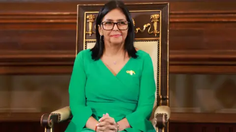 Anadolu via Getty Images Delcy Rodríguez sits on a large chair with her hands clasped in front of her. She wears a plain green dress and large glasses.