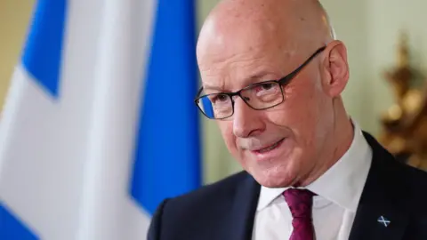 John Swinney in dark suit and white shirt with a burgundy tie in front of a Saltire, speaking at a podium.