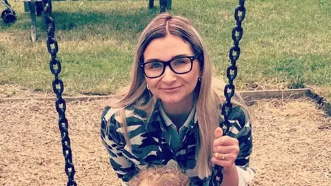 Facebook Isobella Knight with long blonde hair wearing large dark brown glasses. She is wearing a black and gold top and is holding onto the chain of a swing in a park. There is a dried grass landing area behind her and green grass beyond that.