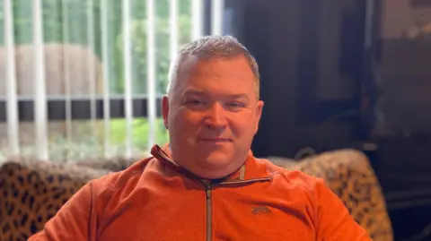 Paul McCaughey sat on a leopard print lounge chair, wearing an orange fleece and looking at the camera