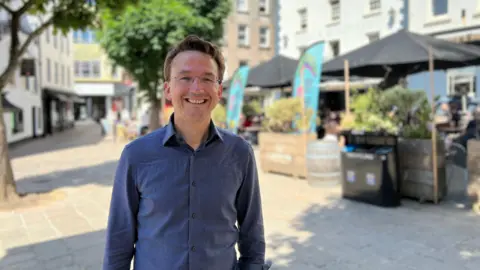 Kirsten Morel wearing a blue button-up shirt stands in an outdoor plaza or street area. The background features buildings, trees, and several tables with umbrellas where people are seated. The sky is clear and blue, suggesting a sunny day.