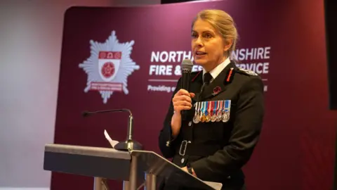 Northamptonshire Fire & Rescue Service Nikki Watson, the chief fire officer, in a black, fire service uniform. There are medals pinned to her jacket. She is standing at a lectern and holding a microphone whilst making a speech. She is in front of a red, branded Northamptonshire Fire & Rescue Service banner.