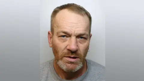 Derbyshire Police A police mug shot of a man with a grey t-shirt, earing, short ginger hair and beard with grey patches.