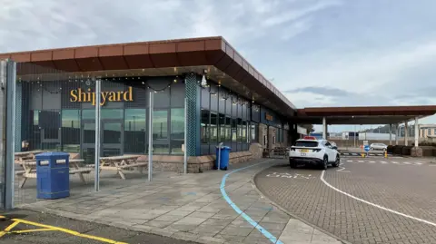 BBC An outside photo of the Shipyard cafe at Elizabeth Harbour in Jersey. There is a white car parked outside. It's a large building with long glass windows. The word shipyard is prominent in yellow writing on the wall. There are benches and a bin behind a metal fence.