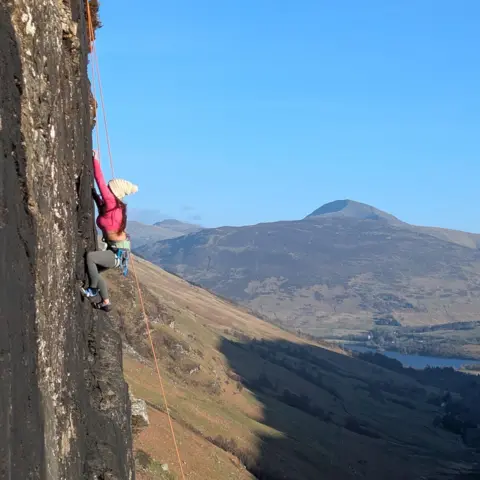 Gathy Tuolor A climber wearing a white bobble hat and red jacket climbs up a steep rock face with ropes. There is a mountain in the distance.