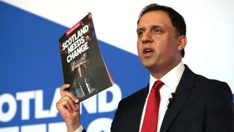Anas Sarwar, who has short black hair, holds up a booklet titles "Scotland needs change". 