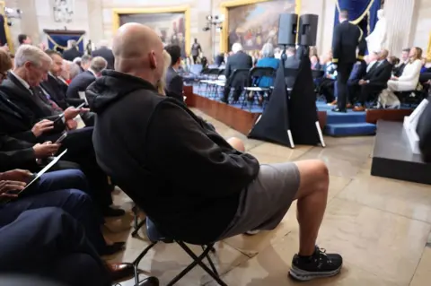 Getty Images A antheral   successful  a hoodie, hoops  shorts and sneakers sits successful  the Capitol Rotunda surrounded by men wearing suits